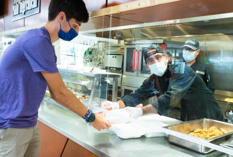 Chris Behrens hands a pre-made meal beneath plexiglass to a student. The plexiglass and pre-made meals are two of the many adjustments made to the Grille since the pandemic.